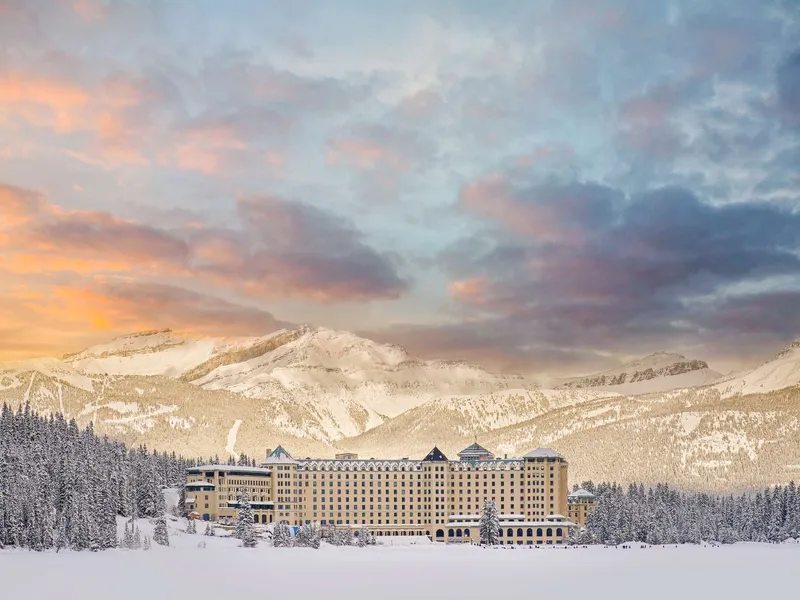 Fairmont Chateau Lake Louise, a stone castle-style hotel, sits on frozen lake with snow-covered mountains and coniferous forest.