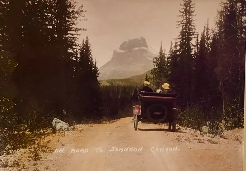 Vintage photograph shows a wagon on the dirt road to Johnston Canyon, with a mountain peak visible through the conifer forest.