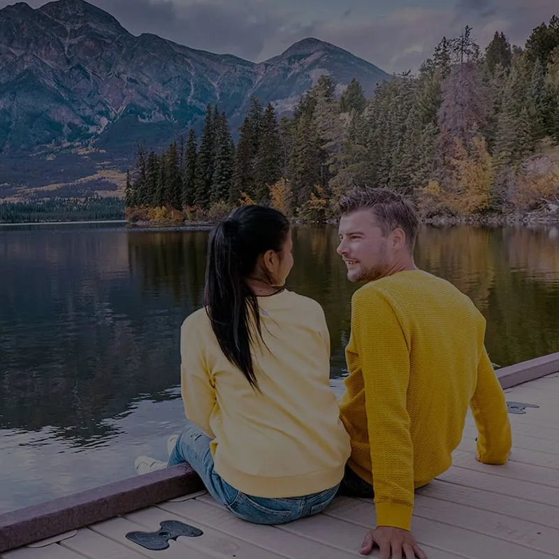 Couple in yellow sweaters sitting on Overlander Lodge's wooden dock overlooking an autumn lake and forested mountains.