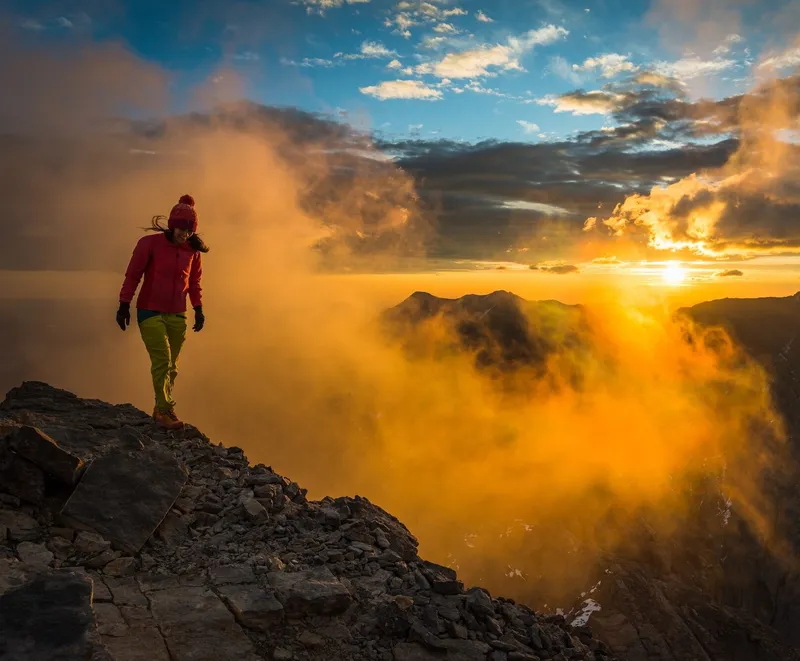 Hiker in red jacket stands on rocky peak above cloud-filled valley at sunrise near Paintbox Lodge.