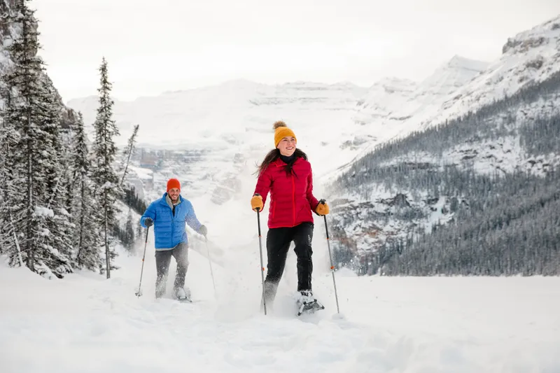 Two skiers in winter gear ascending snowy slopes near Paradise Lodge with snow-capped mountains in background.