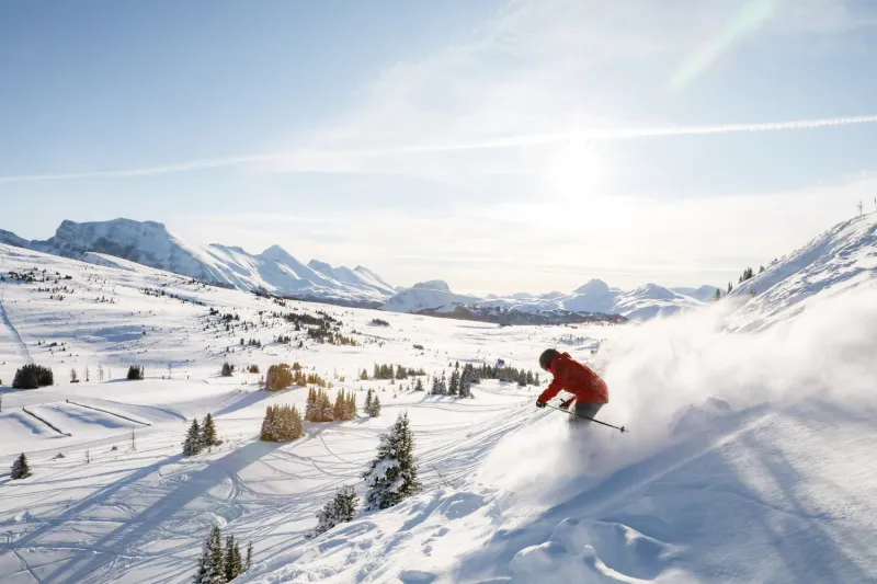 Skier descends snowy slope at Sunshine Mountain Lodge with mountain peaks and forested valley below.